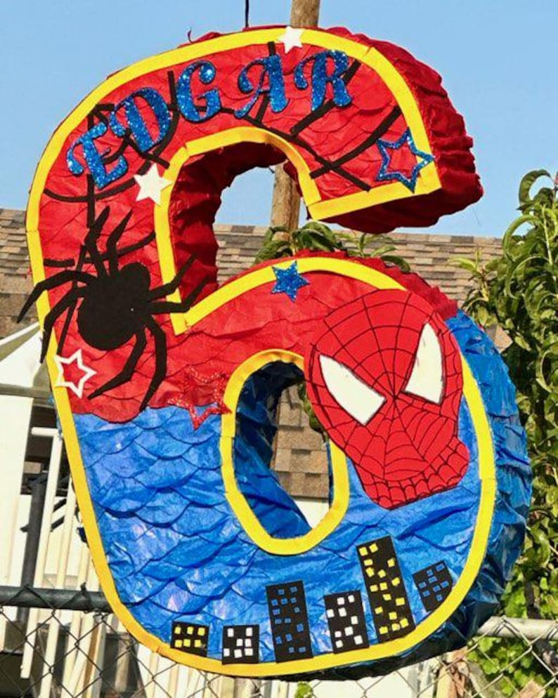 Pu&ograve; includere: Una pignatta a forma di numero sei, rossa, blu e gialla, decorata con un ragno, una maschera di Spider-Man e il nome "EDGAR" in lettere glitterate blu. La pignatta &egrave; a tema supereroi.