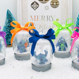 May include: Five glass snow globes with blue Stitch characters inside. Each globe has a different colored ribbon tied around the top. The globes are sitting on a white surface. The text "Merry & Bright" is visible in the background.