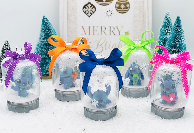 May include: Five glass snow globes with blue Stitch characters inside. Each globe has a different colored ribbon tied around the top. The globes are sitting on a white surface. The text "Merry & Bright" is visible in the background.