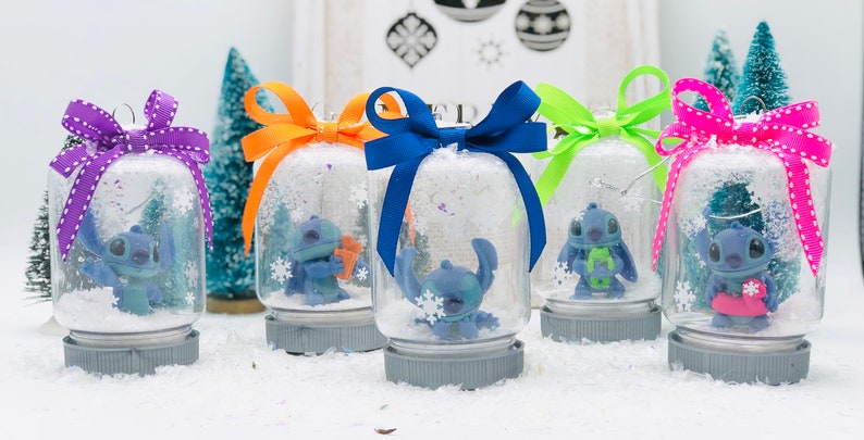 May include: Five snow globes with blue Stitch characters inside. Each globe has a different colored bow on top. The globes are sitting on a white surface.