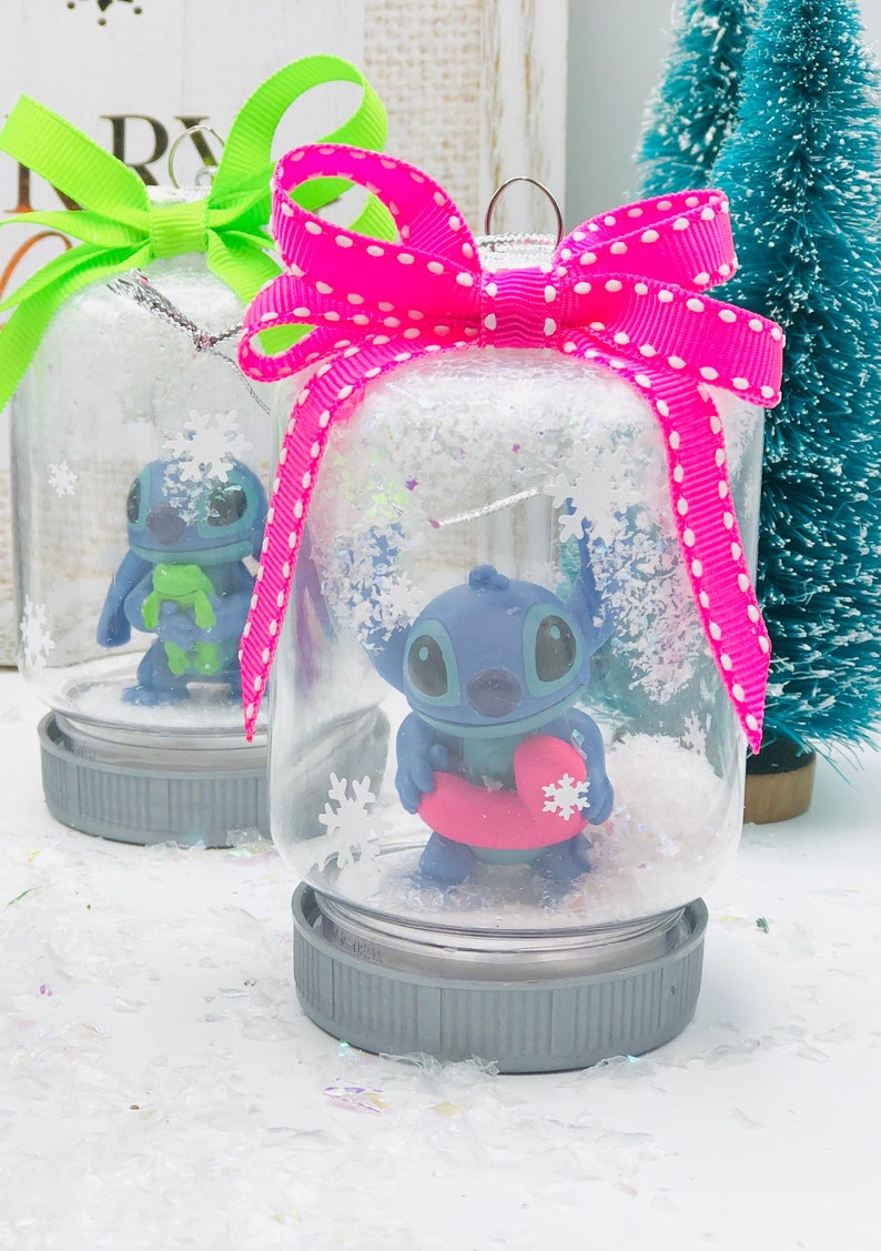 May include: Two glass snow globes with gray lids and pink and green bows. The snow globes contain a blue Stitch character from Lilo & Stitch. One Stitch is holding a green frog and the other is holding a pink inner tube.