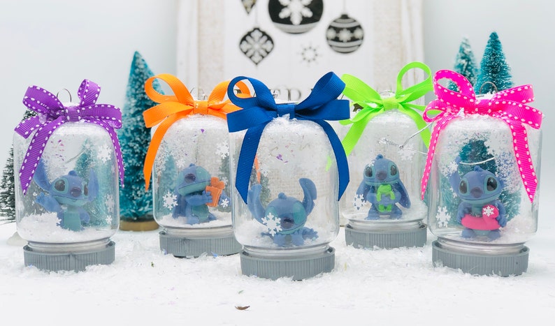 May include: Five snow globes with blue Stitch characters inside. Each globe has a different colored ribbon tied around the top. The globes are sitting on a white surface.