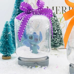 May include: A clear glass snow globe ornament with a purple and white polka dot bow. Inside the globe is a blue Stitch character from the Disney movie Lilo & Stitch. The globe is sitting on a white surface with confetti.
