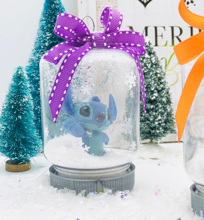 May include: A clear glass snow globe ornament with a purple and white polka dot bow. Inside the globe is a blue Stitch character from the Disney movie Lilo & Stitch. The globe is sitting on a white surface with confetti.