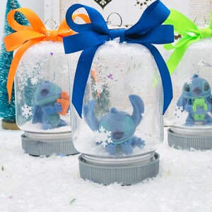 May include: Three glass snow globes with blue, orange, and green ribbon bows. Each globe features a blue Stitch character from the Disney movie Lilo & Stitch. The globes are filled with white glitter and snowflakes.
