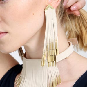 May include: Cream-colored leather fringe earrings and a matching choker necklace with gold accents. The jewelry is worn by a person wearing a navy blue top. The earrings and necklace have a geometric design.
