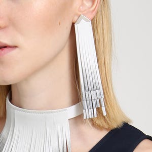 May include: A white leather choker necklace with fringe detail and matching long, fringed earrings. The necklace and earrings have a gradient effect, transitioning from white to silver. The jewelry is worn with a dark blue top.
