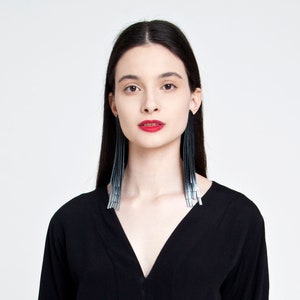 May include: A woman with long black hair wearing a black v-neck top and long silver and black tassel earrings.