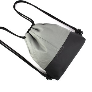 Peut inclure: Un sac à dos gris et noir à cordon avec une finition en cuir. Le sac a un seul compartiment et une fermeture à cordon. Le sac a une étiquette en cuir noir avec le mot "DilliDally" dessus.