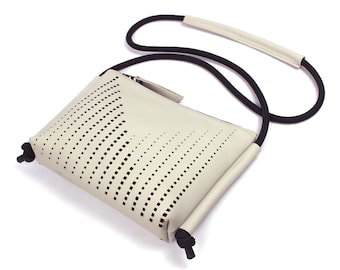 May include: Cream-coloured crossbody bag with a perforated design. The bag features a black rope strap with a cream-coloured shoulder pad. The bag has a rectangular shape with a zip closure and black knotted accents.