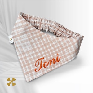 Könnte beinhalten: Ein dreieckiges Haustierhalstuch mit einem hellbeigen und weißen Vichy-Karo-Muster. Der Stoff ist oben mit einem Gummiband gerafft. Der Name Toni ist in orangefarbener Schreibschrift auf der Vorderseite eingestickt. Ein kleines weißes Etikett ist angebracht.