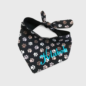 May include: Black dog bandana with a white and brown paw print pattern. The bandana has a turquoise embroidered name "Klabak" on it.