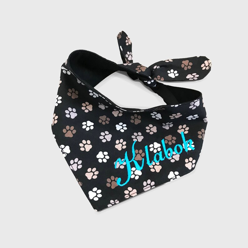 May include: Black dog bandana with a white and brown paw print pattern. The bandana has a turquoise embroidered name "Klabak" on it.