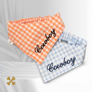 Może przedstawiać: Dwie bandany dla zwierząt w kratkę vichy, jedna pomarańczowa i jedna jasnoniebieska. Każda bandana ma elastyczny pasek na szyję, a z przodu wyhaftowane ciemną nicią słowo Cowboy. Z boku znajduje się mała biała metka.