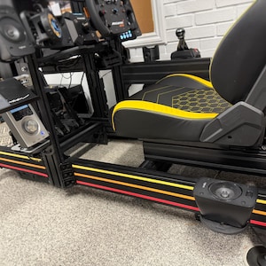 May include: A black racing simulator with a yellow and black racing seat. The simulator has a black metal frame with red, yellow, and black stripes. The seat has a honeycomb pattern and is adjustable. The simulator is on a carpeted floor.