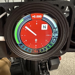 Peut inclure: Un indicateur numérique avec un cadran rouge, affichant la vitesse et d'autres mesures. La jauge a une lunette noire et est montée sur un support noir. L'écran affiche des chiffres et des lettres, dont "0", "N" et "7.9".