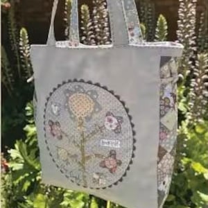 May include: A light gray tote bag with a floral design and the text "Signs of Spring Bag." The bag features a large, circular applique with a flower and butterfly motif. The handles and trim have a patterned design.