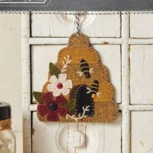 May include: A felt ornament depicting a beehive with a bee and flowers. The ornament is 4 inches by 4 inches and is a craft project from Buttermilk Basin.