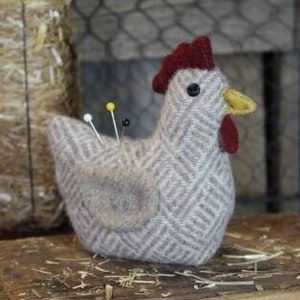 May include: A gray and white patterned chicken pincushion with a red comb and yellow beak. The pincushion is sitting on a wooden surface with the text "#655 - Chicken Pin Keep - 4 1/2" visible.