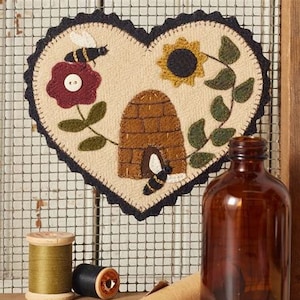 May include: A felt heart-shaped wall hanging with a beehive, sunflower, and red flower with a bee. The beehive is brown and has a small opening. The sunflower is yellow with a black center. The red flower has a button in the center. The bee is black and yellow.