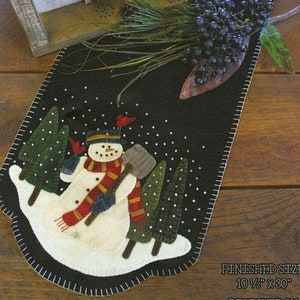 May include: A black felt table runner with a snowman design. The snowman is holding a shovel and wearing a red and yellow scarf. There are small green trees and white snowflakes on the runner. The finished size is 10 1/2 inches by 30 inches.