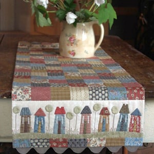 May include: A patchwork table runner with a whimsical village scene. The runner features a grid of various fabric squares in muted colors. The bottom border showcases stylized houses and trees. A floral pitcher with white flowers sits on the runner.