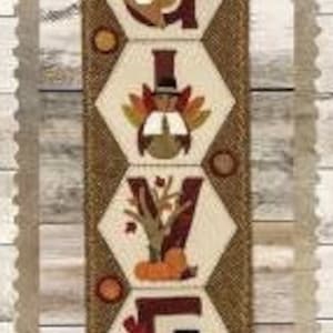 May include: A vertical fabric banner with the word "GIVE" spelled out in brown, beige, and orange fabric. Each letter is decorated with a different Thanksgiving-themed image, including a cornucopia, a pilgrim, a pumpkin, and a turkey.
