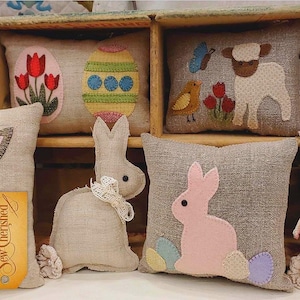 Puede incluir: Una colección de almohadas de Pascua hechas a mano con conejos, un cordero y huevos de Pascua. Las almohadas están hechas de tela de arpillera y tienen un estilo rústico y vintage. Las almohadas están decoradas con detalles bordados y tienen una etiqueta "Sew Cherished" adjunta.