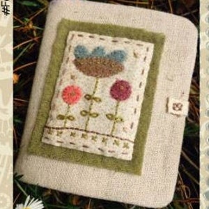 May include: A small, beige linen notebook with a green felt cover. The cover features an embroidered design of three flowers in shades of brown, blue, pink, and green.