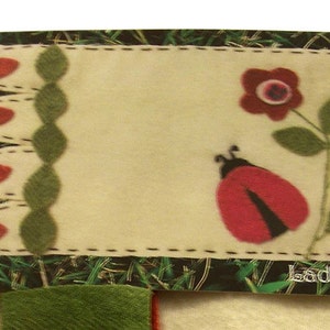 May include: A white felt placemat with a red ladybug, a red flower, and green leaves. The placemat has a stitched border with red and green accents. The text "ladybug" is printed in the bottom right corner.