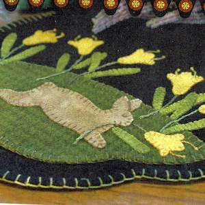 May include: A brown rabbit peeking out from behind a green bush with yellow flowers. The rabbit is made of felt and is surrounded by a black background with a green border.
