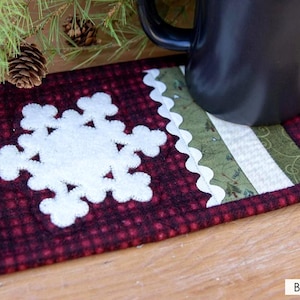 May include: A burgundy and black plaid fabric coaster with a white snowflake applique. The coaster has a green and white patterned section and a scalloped edge. A black mug sits on the coaster, with pine needles and pine cones in the background.