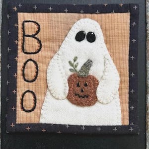 May include: A Halloween-themed fabric art piece featuring a white ghost holding a brown pumpkin. The word "BOO" is stitched in black thread. The artwork is framed with a dark blue border with cross stitch details.