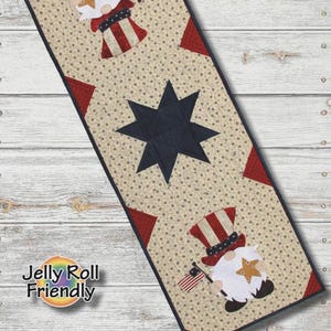 Op de afbeelding: Een rechthoekig tafelloper quiltpatroon met een beige achtergrond met kleine donkere sterren, een grote centrale marineblauwe ster en twee kabouters met rood-wit gestreepte hoeden die kleine vlaggetjes en gouden sterren vasthouden.