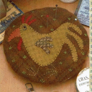 May include: A brown felt pincushion shaped like a chicken with a red comb and wattle. The chicken is surrounded by green felt grass and brown felt stitching.