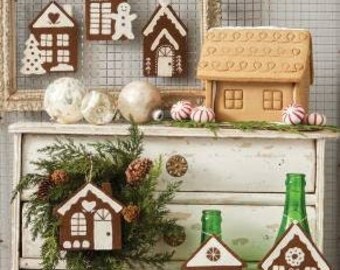 Peut inclure: Une collection d'ornements en forme de maison en pain d'épices et une plus grande maison en pain d'épices. Les ornements sont brun et blanc avec différents motifs, dont un bonhomme de neige, un arbre et un cœur. La plus grande maison en pain d'épices est brune et a une porte et des fenêtres blanches.