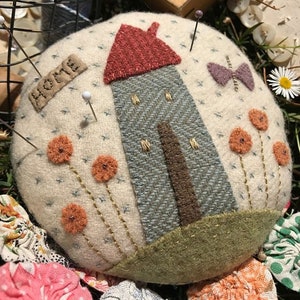 May include: A white felt pincushion with a house, flowers, and a butterfly embroidered on it. The house has a red roof and a brown door. The flowers are orange and the butterfly is purple. The pincushion has the word "HOME" embroidered on it.
