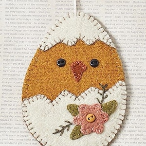 Puede incluir: Un adorno de fieltro con forma de pollito con cuerpo marrón, cáscara blanca y una flor rosa. El pollito tiene ojos negros y un pico rojo.