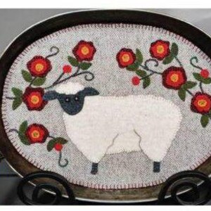 Puede incluir: Una oveja blanca con ojos negros y una nariz negra está rodeada de flores rojas sobre una alfombra ovalada gris. La alfombra está en una bandeja de metal con un asa negra.