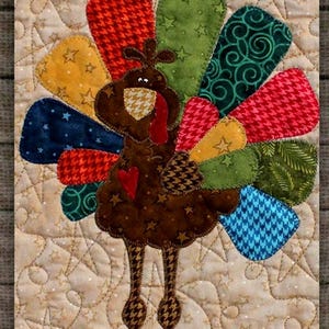 May include: A quilted Thanksgiving turkey with a brown body, patterned legs, and a colorful fan of feathers. The feathers feature various patterns and colors, including red houndstooth, green stars, and blue geometric designs. The background is a beige fabric with star-shaped quilting.