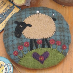 May include: A round pincushion with a felt sheep design. The sheep is brown and white with black eyes and a black nose. The sheep is standing on a green hill with red flowers. There is a purple heart on the hill. The pincushion is made of felt and has a blue and green background.