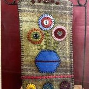 May include: A felt wall hanging featuring a blue vase and colourful flowers. The flowers are made with felt circles and buttons. The vase is outlined with brown stitching.