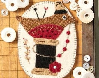 May include: A brown and red felt bird with a black hat and a spool of thread for a body. The bird is perched on a spool of thread with a red flower at the base.