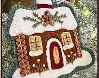 Peut inclure: Une décoration de maison en pain d'épices en feutre avec un bonhomme en pain d'épices, une porte rouge et de la neige blanche. La maison est décorée de feuilles vertes et d'une cheminée rouge.