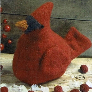 May include: A red felt cardinal bird ornament with a black beak and a brown felt beak. The ornament is sitting on a wooden surface with white flakes and red berries scattered around it.