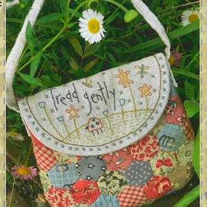 Op de afbeelding: Handgemaakte patchwork crossbodytas met een crèmekleurige riem. De flap heeft de tekst "Tread gently" en geborduurde bloemen. De tas is gemaakt van verschillende stoffen met patronen in rood, blauw en grijs.