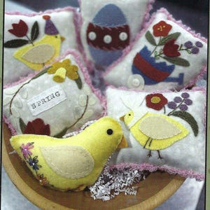 May include: A set of five felt pillows with spring-themed embroidery. The pillows feature a yellow chick, a blue egg, a red flower, a yellow chick, and a red flower. The pillows are all decorated with pink lace trim. The pillows are arranged in a wooden bowl.