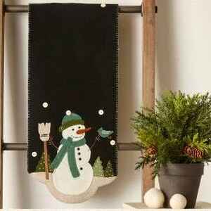 May include: Black felt table runner with a snowman design. The snowman is wearing a green scarf and hat and holding a blue bird. The background is a dark green with white snowflakes.