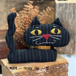 May include: A black Crazy Cat plush toy, approximately 5 inches tall, with yellow eyes, a red nose, and a gold collar. The cat has a stitched white mouth and whiskers. The text "cottonwood creations" and "designer jennifer clemen" are at the top.