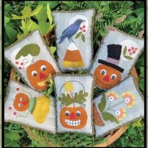 May include: Six handmade felt pumpkin pillow ornaments in a basket. The pumpkins are decorated with different designs, including a crow, a candy corn, a top hat, a flower, a bucket, and a bug.